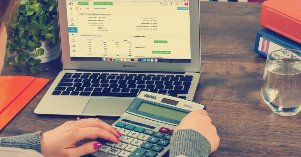 Hands working out care costs on a calculator beside a laptop showing an account statement at a wooden desk
