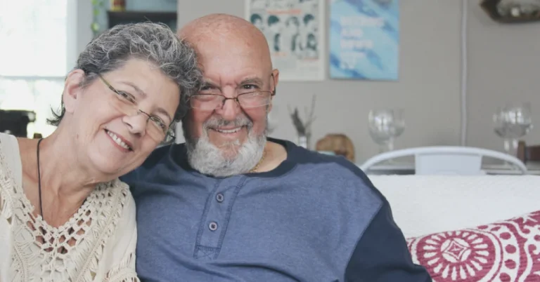 An older couple sit together on a sofa in their own living room, smiling, the setting a family care manager would visit and assess.