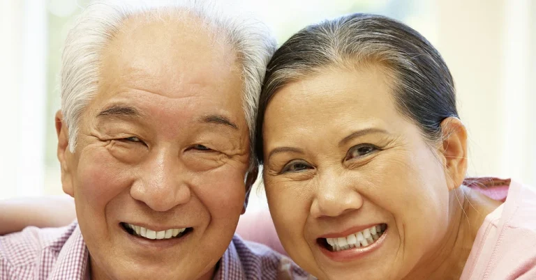 An older couple sit close together and smile in a bright room at home, the kind of independent living a medical alert system supports.