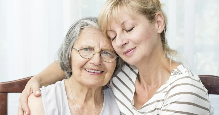 A smiling older woman in glasses rests against her adult daughter, who has an arm around her shoulders in a bright room at home.