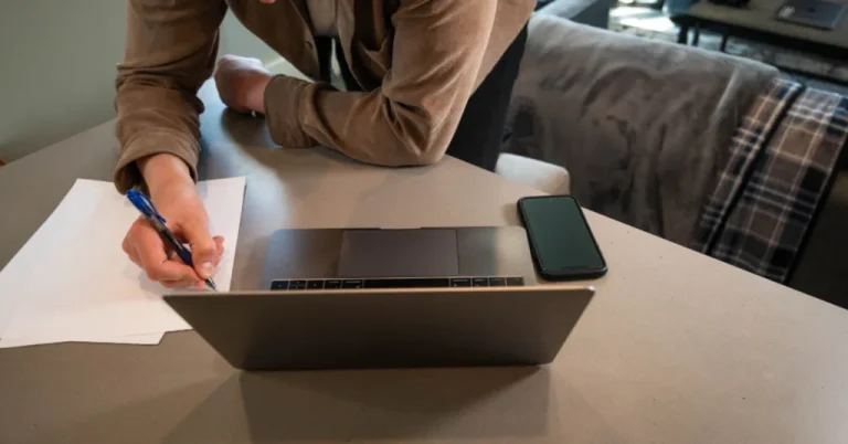 A person writes notes on paper next to an open laptop and phone at a home desk, managing caregiving tasks.